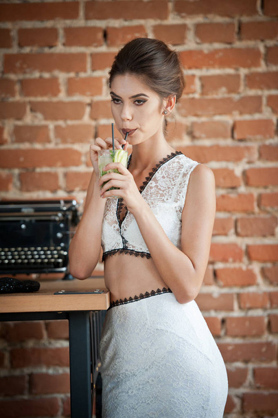 Fashionable attractive lady with white dress  standing near a restaurant table having a drink. Short hair brunette woman with makeup and creative haircut holding a glass with fresh - Foto, imagen