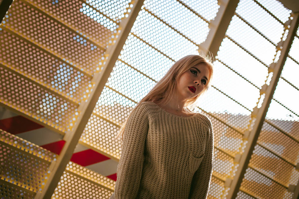 Portrait of a young woman dressed in a knitted sweater and blue jeans. Standing near a metal perforated parking fence construction - Photo, Image