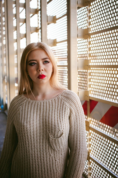 Portrait of a young woman dressed in a knitted sweater and blue jeans. Standing near a metal perforated parking fence construction - Fotoğraf, Görsel