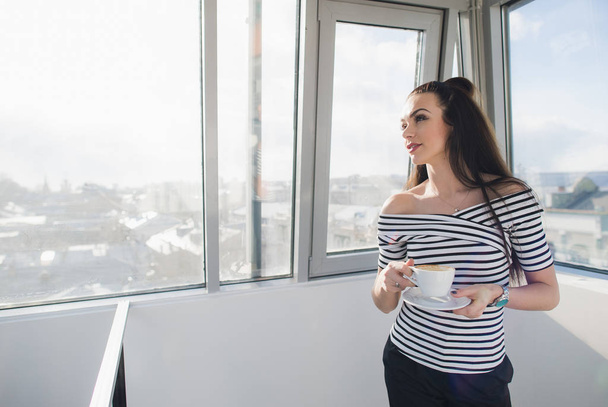 Портрет дорослої красивої жінки, яка дивиться через вікно, стоячи з чашкою кави
. - Фото, зображення