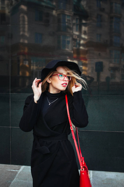 Beautiful young woman in a black coat and hat, wearing red glasses and a red bag walking in the city. Reflection of the city in the window - Foto, imagen