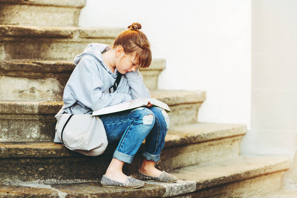 Dziecko dziewczynka czytanie książki poza, siedząc na schodach, noszenie szara Bluza i plecak - Zdjęcie, obraz