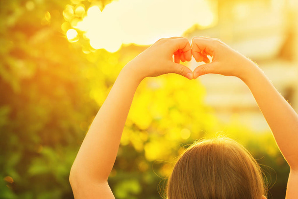Kislány, így a szív jel vele kezet a nyári kertben naplementekor  - Fotó, kép