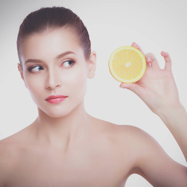 Beautiful young shirtless woman holding piece of orange in front of her eye while standing against white background - Photo, Image
