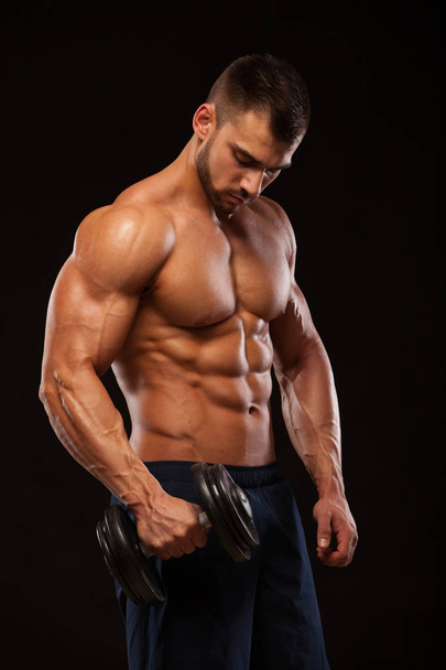 Handsome power athletic man with dumbbell is confidently looking forward. Strong bodybuilder with six pack, perfect abs, shoulders, biceps, triceps and chest Isolated on black background with - Foto, imagen