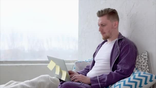 Uomo con computer portatile seduto a letto comunica con gli amici nei social network
. - Filmati, video