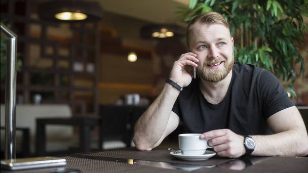 Young bearded attractive businessman in blue shirt is sitting at wooden table in restaurant, talking on phone and drinking tea. Man is waiting for colleague in cafe. Blurred background. Lifestyle. - Photo, image