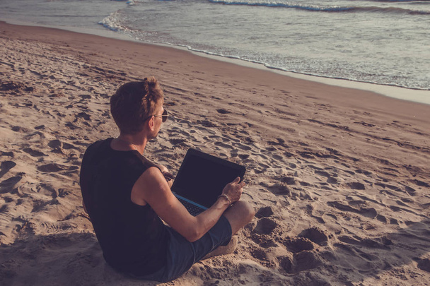 Nuori mies työskentelee kannettavan tietokoneen parissa rannalla. Vähennys
 - Valokuva, kuva