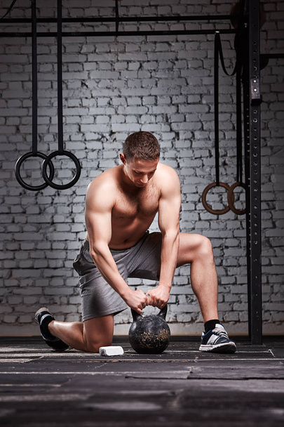 Young sportsman holding kettlebell with his arms on the cross fit gym floor against brick wall. - 写真・画像