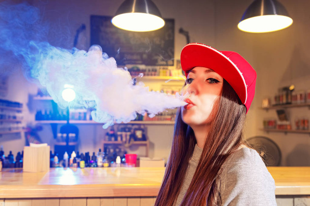 Jovencita guapa con gorra roja fuma un cigarrillo electrónico en la tienda de vapores
 - Foto, imagen