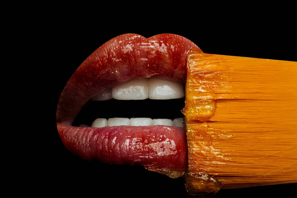 Концепция косметики Lip. Блеск для губ выделен на черном фоне, женский рот с красными губами и зубами. Кисть для помады и блеск близко. Чувственный женский рот и поцелуй. Французский поцелуй
 - Фото, изображение