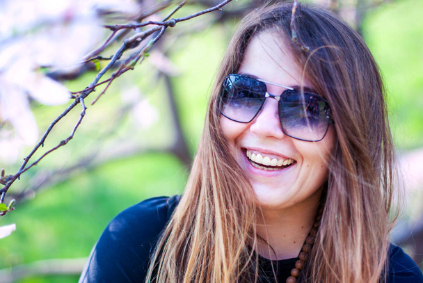 Bella ragazza in occhiali da sole sorridente vicino alberi in fiore. La cicciona sorride nel giardino della magnolia. Ragazza con i capelli lunghi che indossa occhiali da sole
. - Foto, immagini