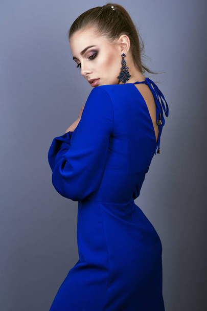 Portrait of young gorgeous model with ponytail and artistic make-up wearing electric blue open back dress and long delicate earrings keeping her hands at her breast - Photo, Image