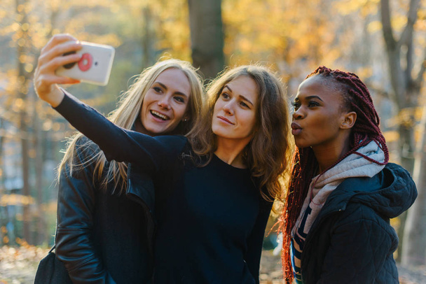 Three attractive happy woman making selfie in autumn park. Pretty girls with different colored skin. Female making funny faces - Fotó, kép