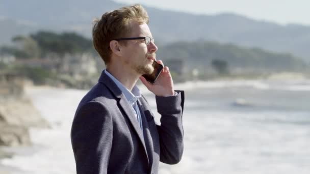 Young businessman stands by the Pacific ocean coast and talks on phone - Video, Çekim