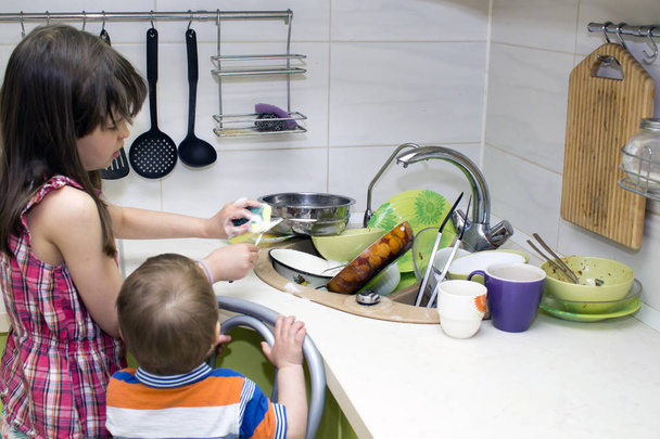 das Kind wäscht ein Bündel schmutziges Geschirr, das in der Nähe der Spüle steht - Foto, Bild