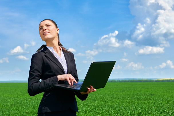 Бизнес-концепция - женщина в зеленом травяном поле наружная работа на ноутбуке. Молодая девушка в черном костюме. Прекрасный весенний пейзаж с облачным небом, яркий солнечный день
 - Фото, изображение