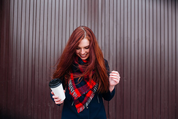 Beautiful happy young woman holding a paper cup and drinking coffee. Cheerful woman with red long hair in the street drinking morning coffee - Φωτογραφία, εικόνα