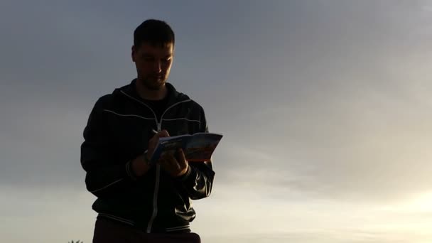 herrlicher Sonnenuntergang und ein junger Mann, der an einer steinigen Küste im Herbst etwas in sein Notizbuch schreibt - Filmmaterial, Video