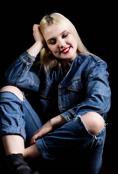Fashion studio photo of young woman in danim jacket and pants with torn knees sitting on floor, dark background. - Fotografie, Obrázek