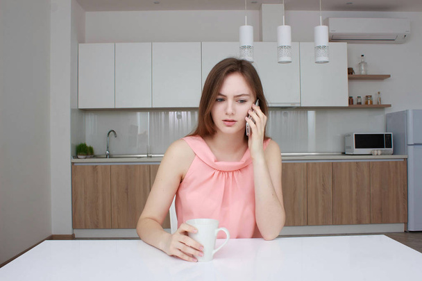Beautiful girl is talking on the mobile phone, holding a cup. kitchen. - Foto, immagini