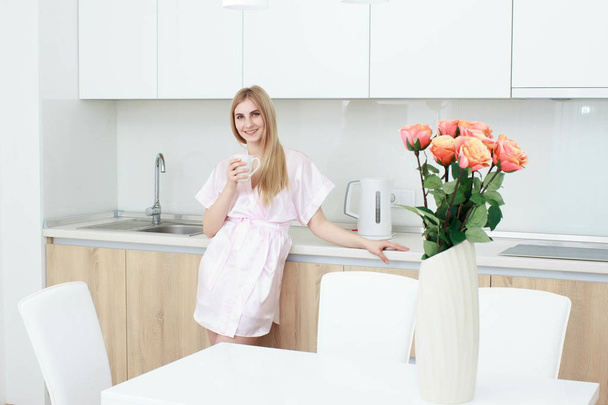 Smiling woman in bathrobe standing with coffee cup at home in the kitchen - Fotografie, Obrázek
