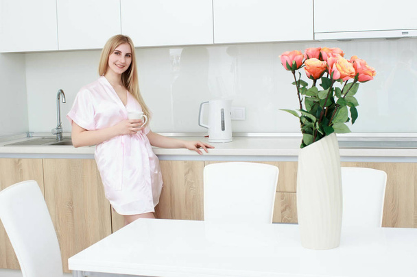 Smiling woman in bathrobe standing with coffee cup at home in the kitchen - Foto, imagen