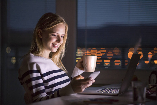 Woman at desk, holding smartphone, working on laptop at night. - Fotografie, Obrázek