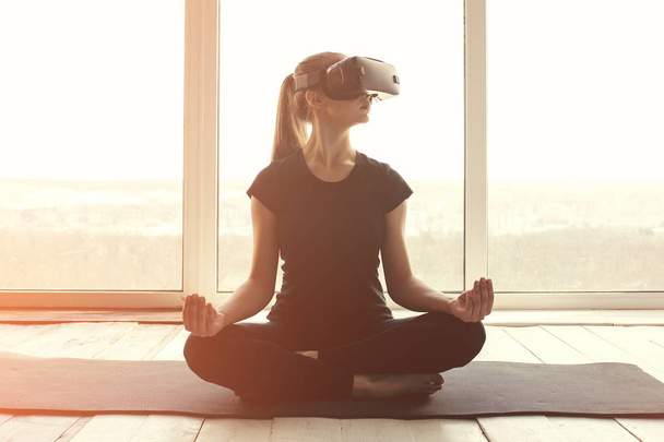 Uma jovem menina bonita em óculos de realidade virtual faz ioga e aeróbica remotamente. Conceito de tecnologia futura. Tecnologia de imagem moderna. Aulas de esportes individuais remotamente
. - Foto, Imagem