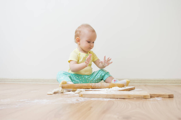 A little cute baby-girl with on her head is sitting on the floor, she is heavily soiled with wheat flour - Zdjęcie, obraz