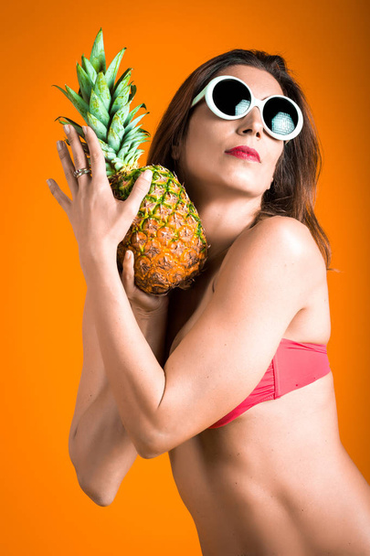 Portrait of young healthy beautiful woman in red bikini and white sunglasses with fresh pineapple in a hand. - Foto, immagini