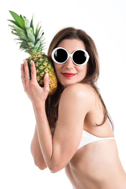 Portrait of young healthy beautiful woman in white bikini and white sunglasses with fresh pineapple in a hand - Fotó, kép
