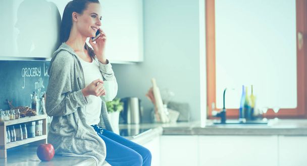 Woman using mobile phone sitting in modern kitchen - Photo, Image