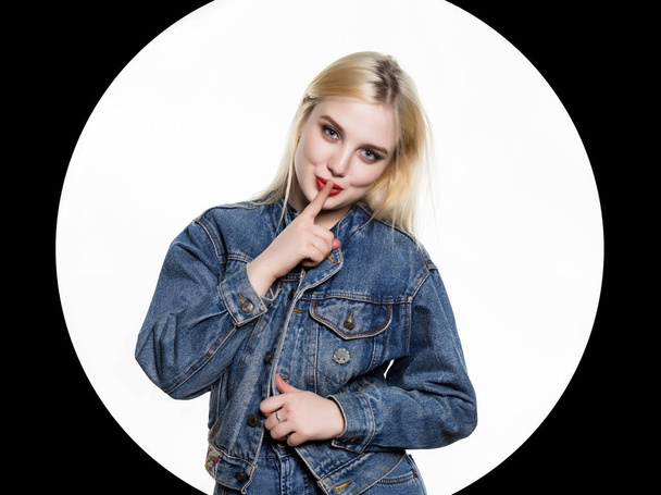 young blonde woman in denim jaket and jeans shows Index finger to the lips on white background - Fotoğraf, Görsel