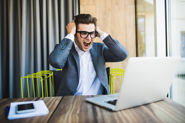 Happy businessman in glasses triumphing with raised fists and screaming look at laptop - Foto, Imagen
