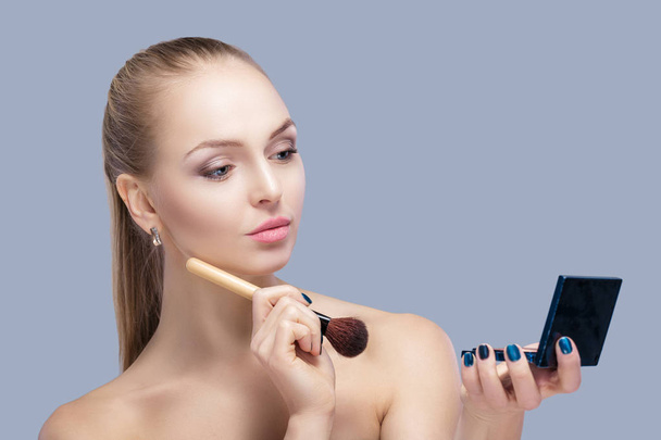 beautiful blond woman holding makeup brush and looking in the mirror on a gray background. - Photo, image