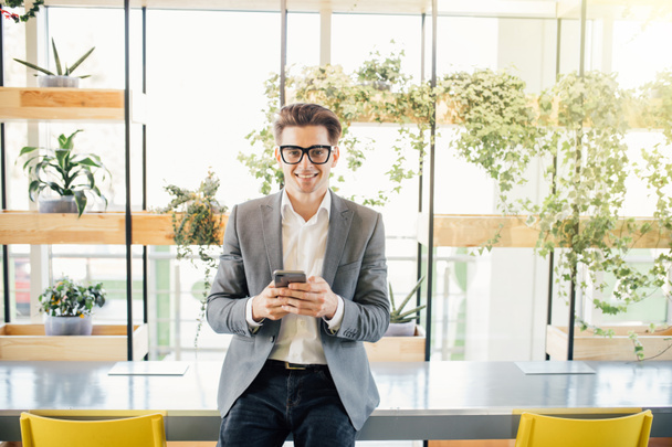 Красивий позаштатний бізнесмен сидить на столі з телефоном в руках, кріпиться в мережі, перевіряє електронну пошту або соціальні мережі в сучасному офісі
 - Фото, зображення