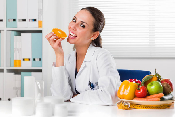 Nainen ravitsemusterapeutti syö Oranssi viipale toimistossaan
 - Valokuva, kuva