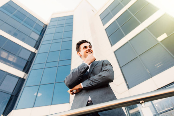 Привлекательный молодой бизнесмен с руками на подбородке перед современным офисным зданием в прекрасный солнечный день
. - Фото, изображение