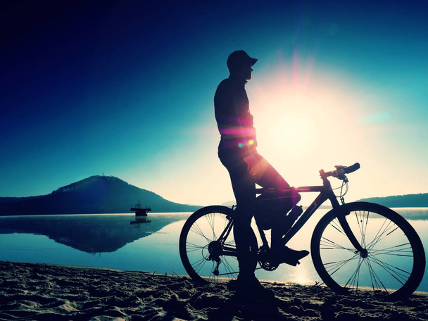 Силуэт спортсмена, держащего велосипед на берегу озера, красочное облачное небо заката на заднем плане
 - Фото, изображение