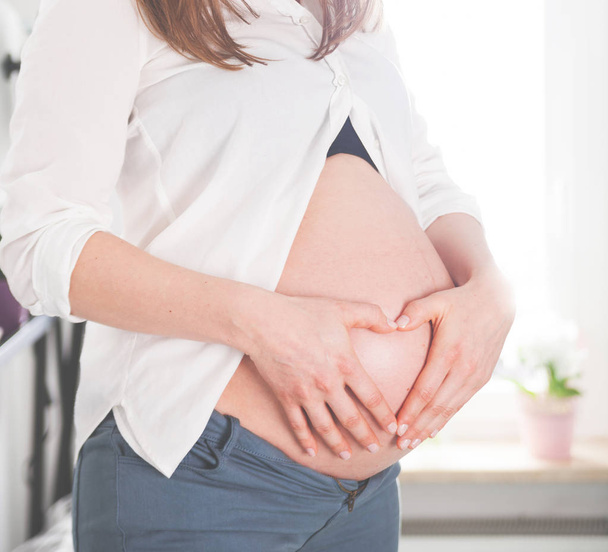 Pregnant girl holding hands in heart shape on belly at home - Fotografie, Obrázek