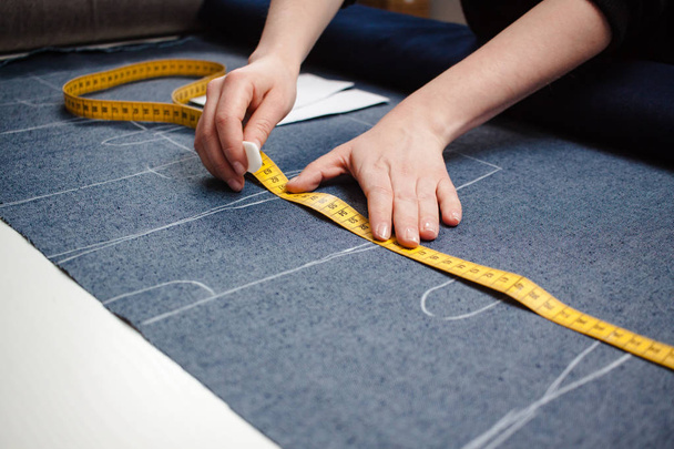 Hands of seamstress cutting a jeans fabric with scissors pattern and measuring tape on white table - Фото, зображення
