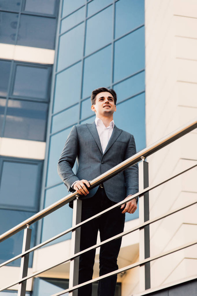 Attractive young confident businessman in classic suit is looking away, leaning on balcony, outside the office building - Foto, Imagem