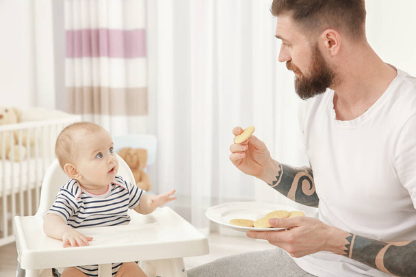 Handsome tattooed young man feeding cute little baby at home - Fotoğraf, Görsel