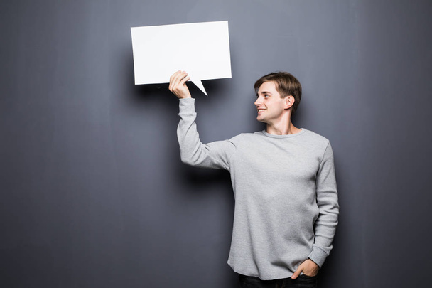 Young man holding white blank speech bubble with space for text look away isolated on grey background. - Foto, Imagen