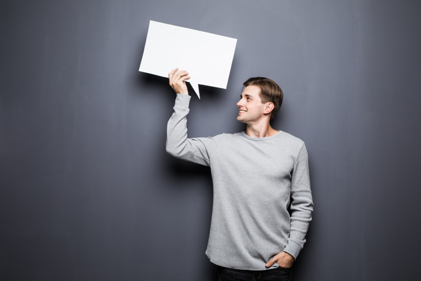 Young man holding white blank speech bubble with space for text and look at it isolated on grey background. - Foto, afbeelding