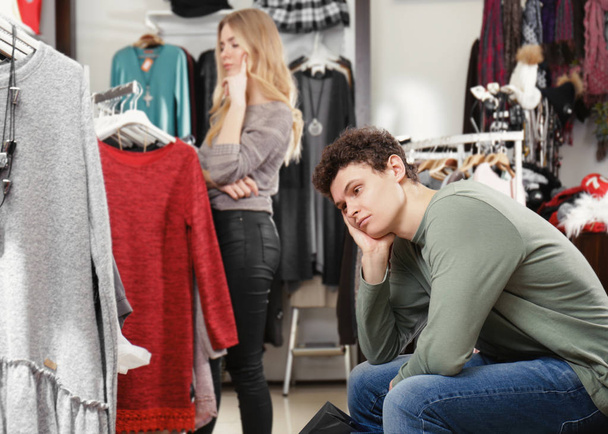 Man waiting his girlfriend while she choosing clothes in modern shop - Photo, image
