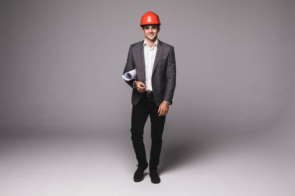 Handsome young architect in suit and protective helmet is holding rolled paper on gray background - Fotoğraf, Görsel