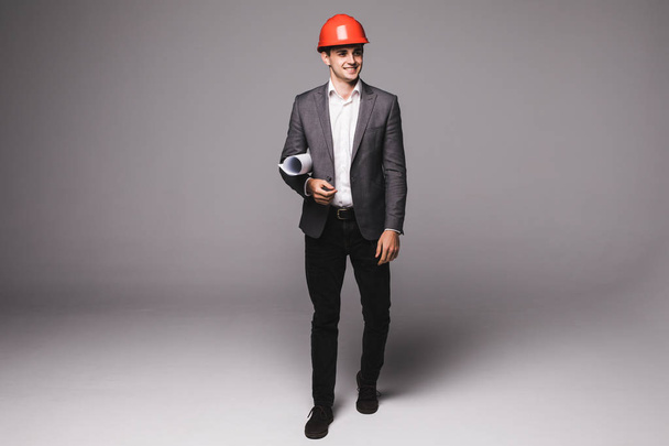 Bonito jovem arquiteto de terno e capacete de proteção está segurando papel laminado em fundo cinza
 - Foto, Imagem
