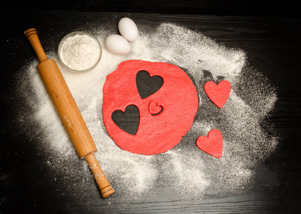 Red hearts cut out dough with rolling pin, eggs and flour on a black table . View from above. Space for text - Photo, Image